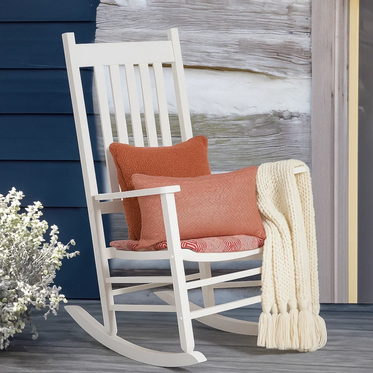 Fauteuil berçant blanc avec des coussins orange et une couverture tricotée crème sur une véranda rustique en bois.