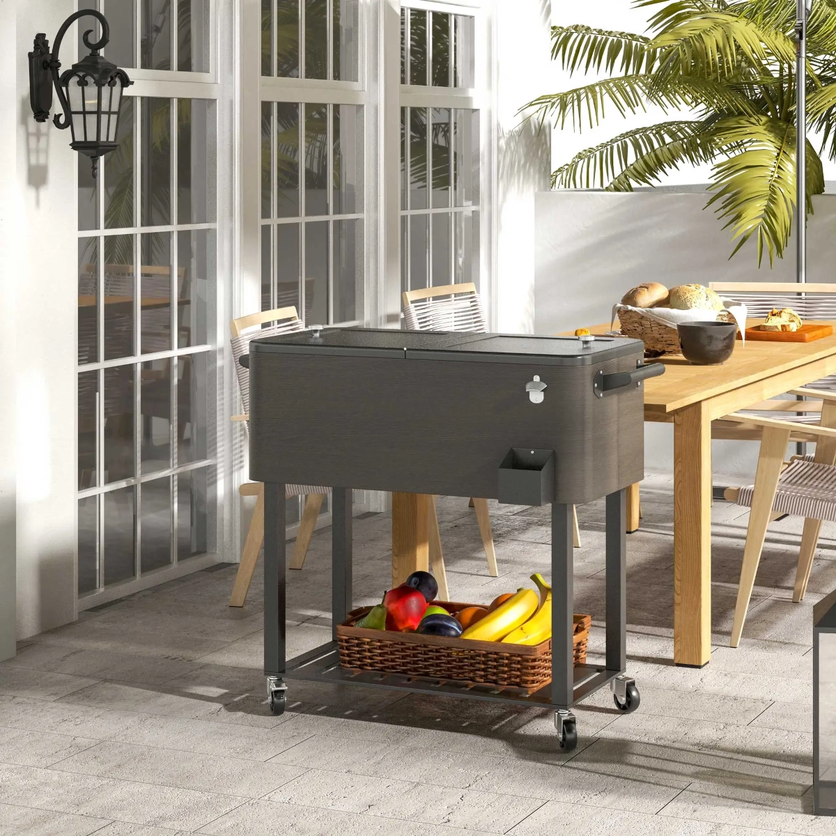 Caddie de boissons à roulettes en extérieur avec un panier de fruits en dessous, placé sur une terrasse à côté d'une table de repas.