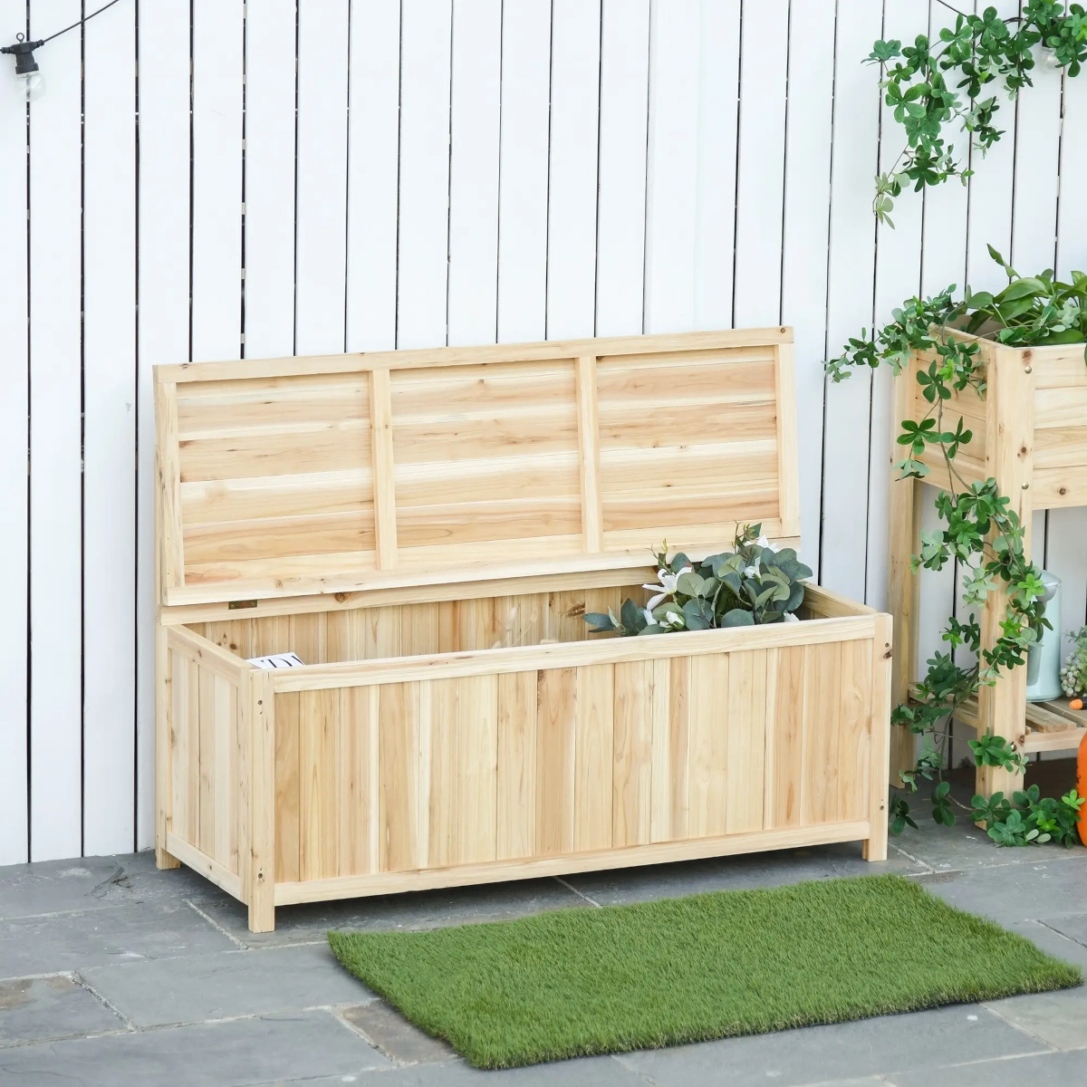 Un banc de rangement en bois naturel avec couvercle ouvert placé contre une clôture blanche dans un jardin.