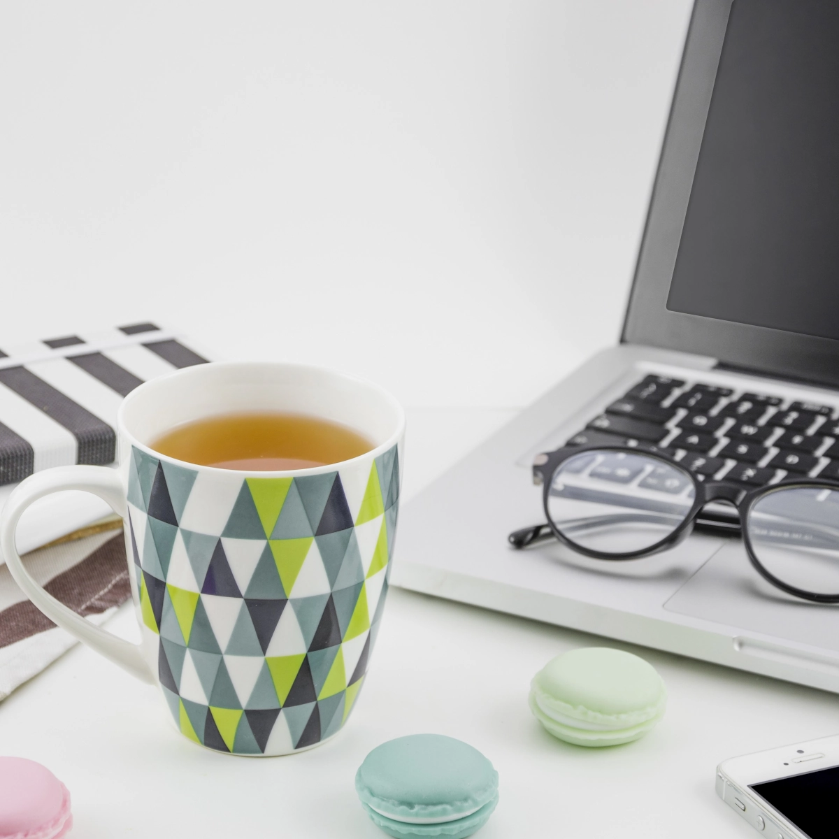 Tasse à motifs avec du thé près d'un ordinateur portable, des macarons, un carnet et des lunettes