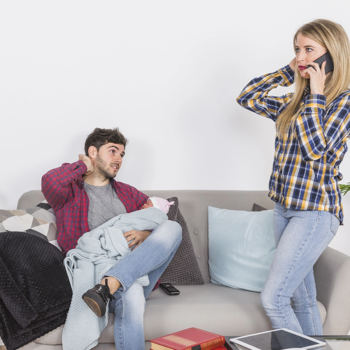 Homme tenant un bébé sur un canapé l'air fatigué, femme parlant au téléphone en arrière-plan.