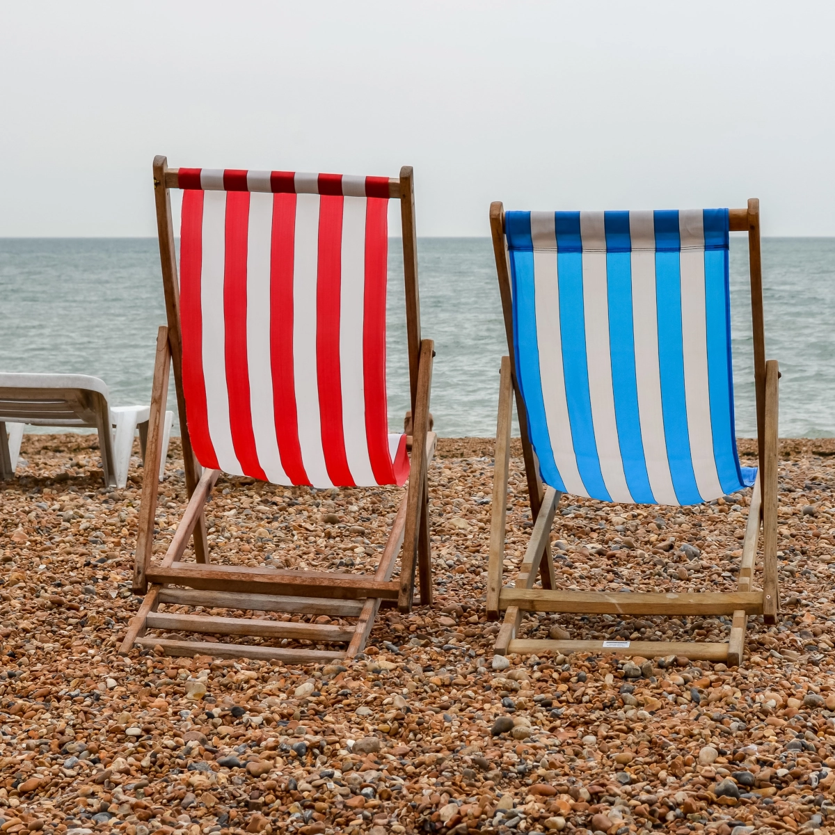 Chaises de plage en bois rayées rouge et bleu face à l'océan sur une plage de galets.